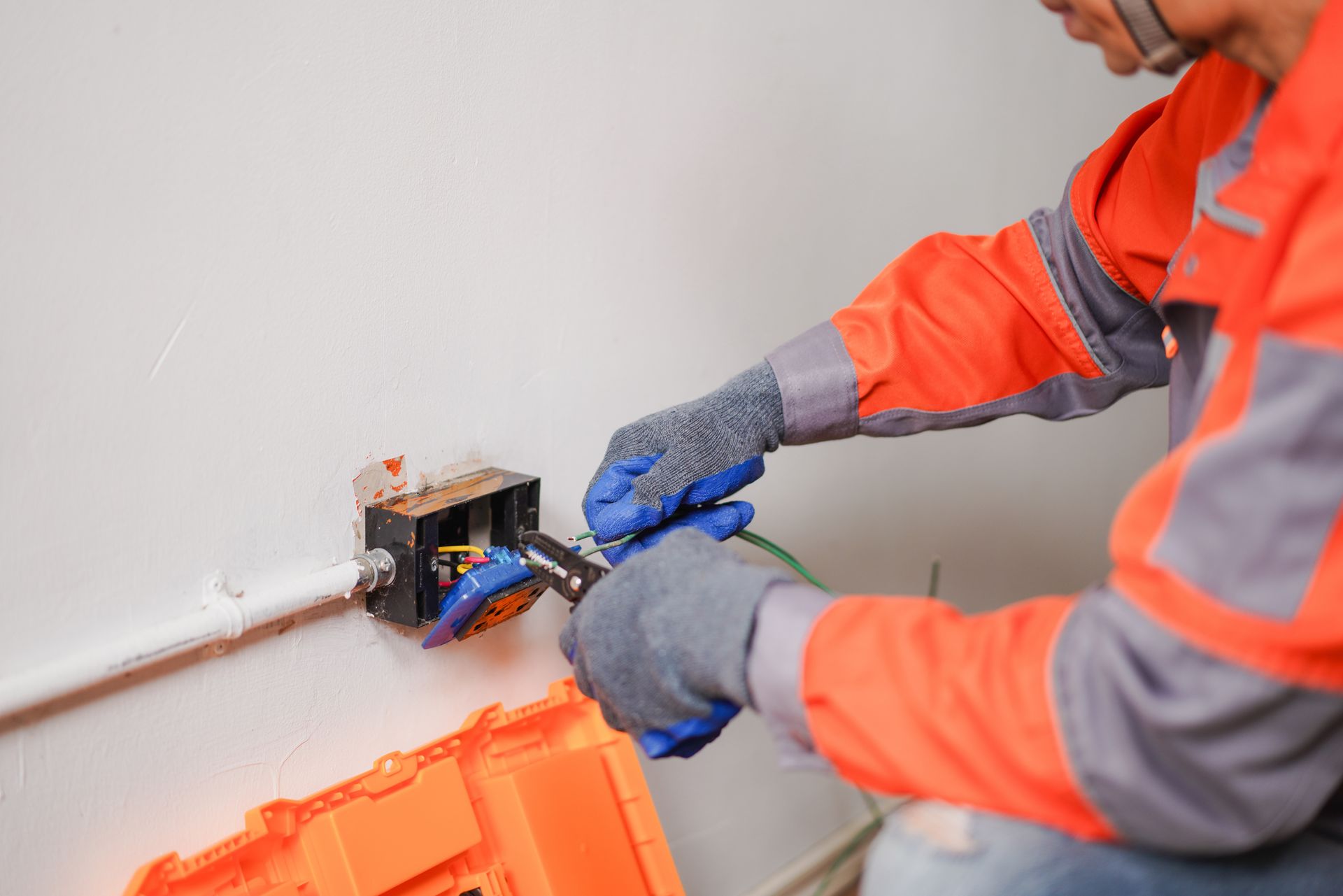 Electrician in orange jacket wiring an outlet with blue gloves and pliers