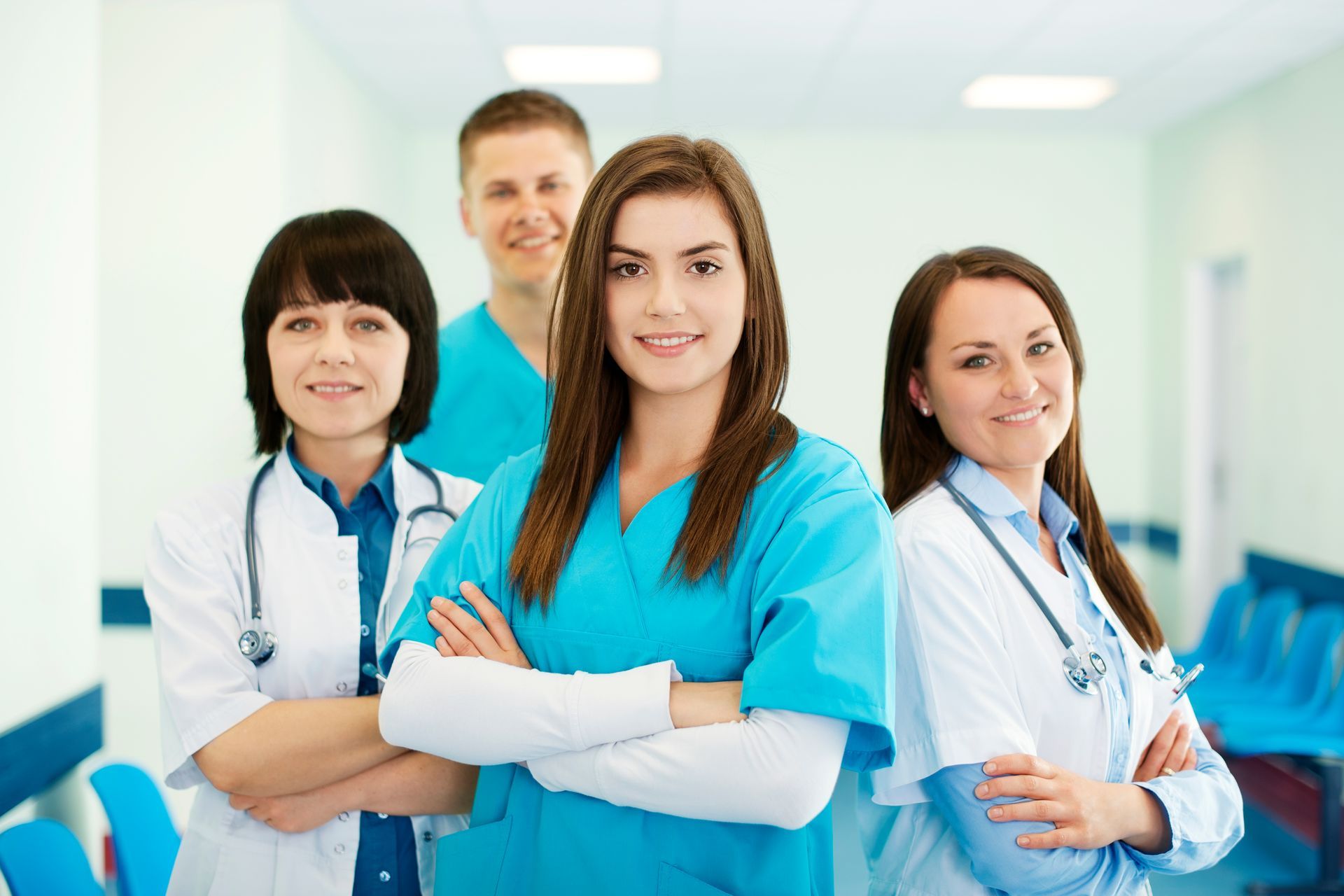 Medical professionals in scrubs and white coats stand in a hospital hallway, arms crossed, smiling at the camera.