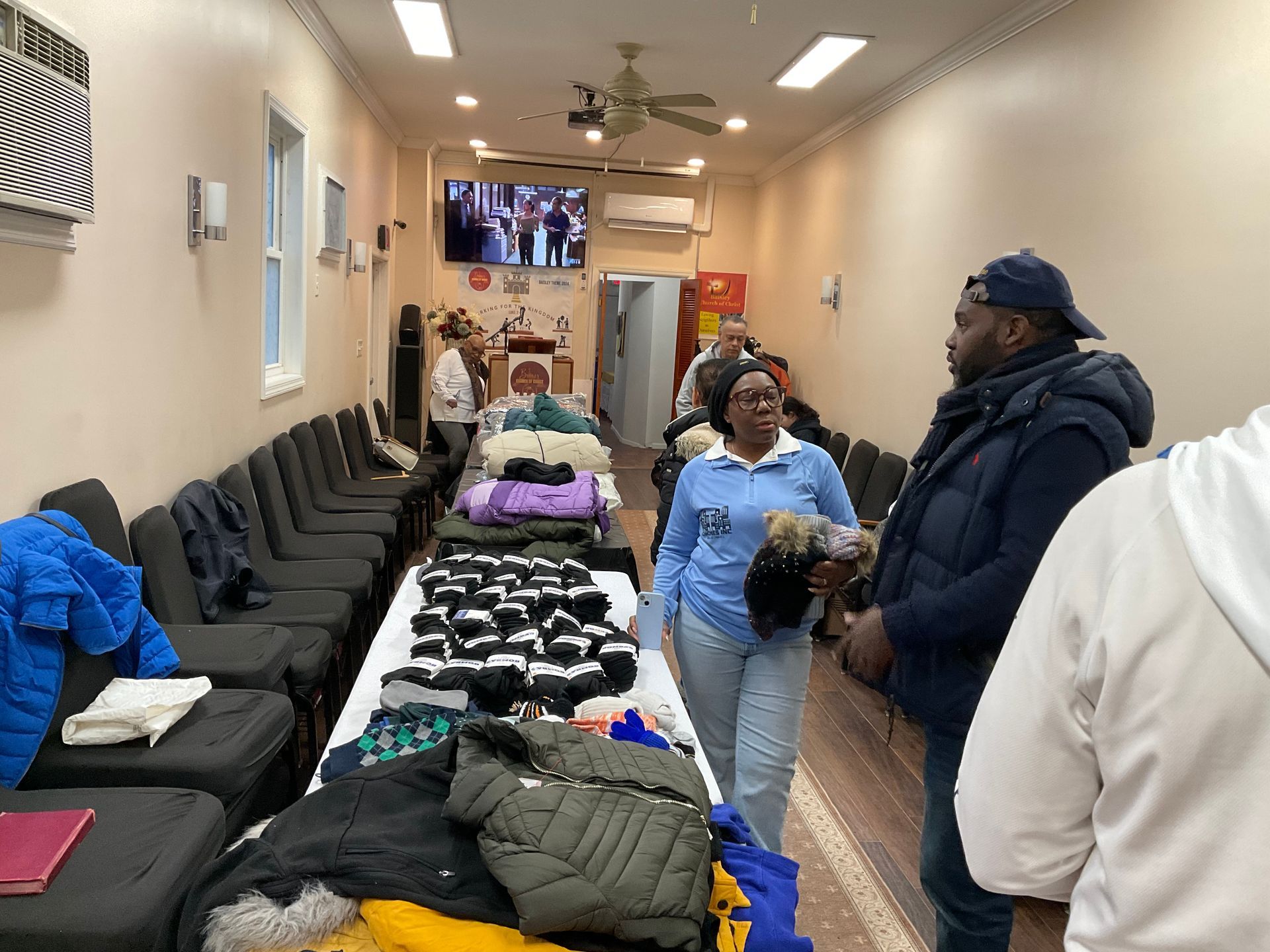 People browse clothing laid out on tables in a long room with seating, likely a donation event.