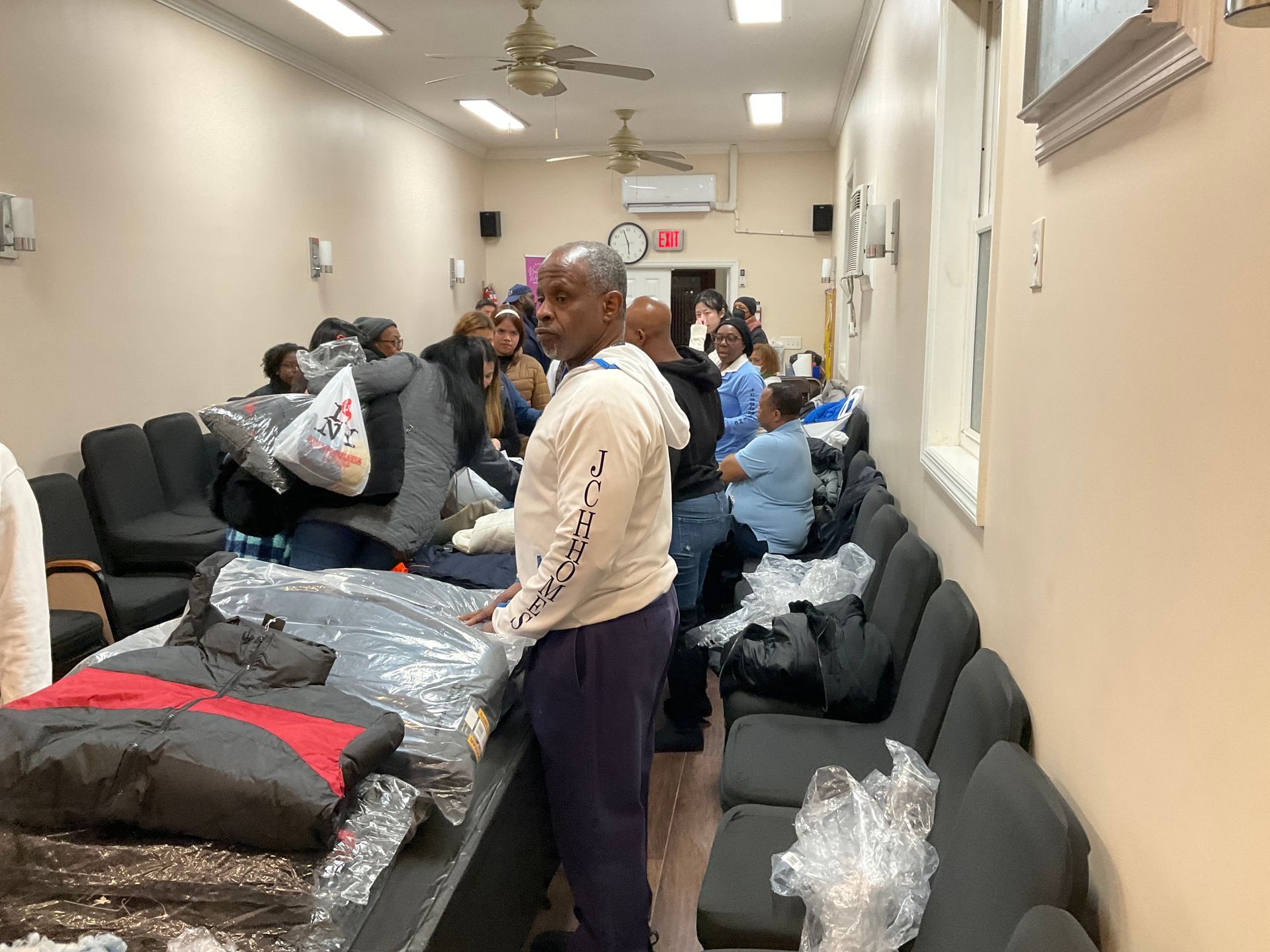 People inside a room; coats on a table. Man in sweatshirt looks at the camera; others in the background.