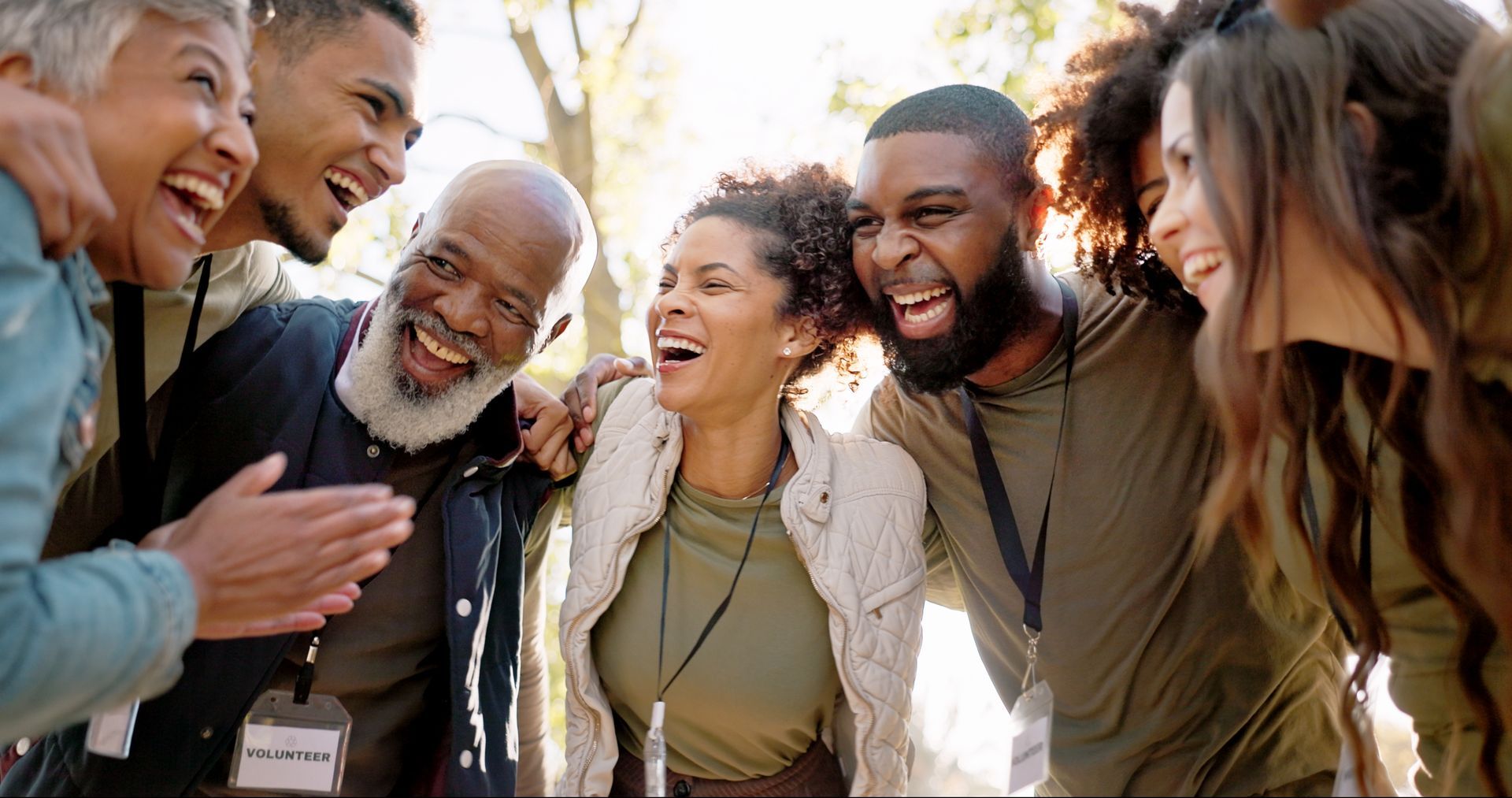 Group of people with various skin tones laughing, embracing outdoors, looking joyful.