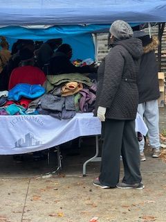 People browsing clothing at an outdoor table. A blue tarp provides overhead cover.