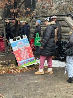 Group of people on a city sidewalk; leaning sign, one person wears winter coat and pink pants.