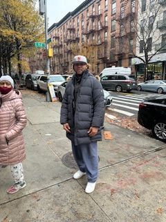 Man in a puffer coat and cap stands on a sidewalk in front of buildings, cars, and a crosswalk.