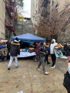People gather around a table under a blue canopy in an urban alley.