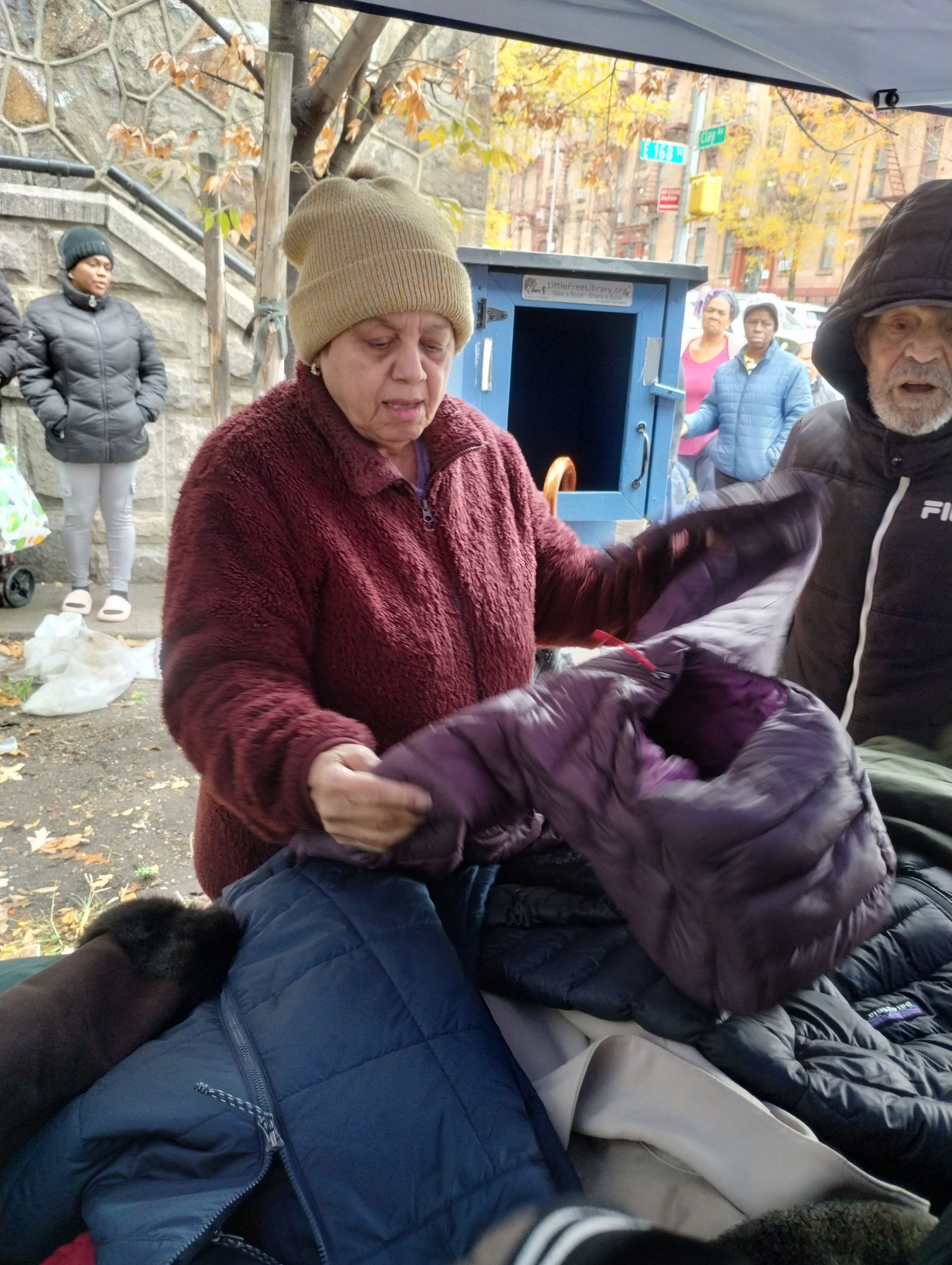 Woman at a table holding a purple coat; outdoor setting with other coats and people.