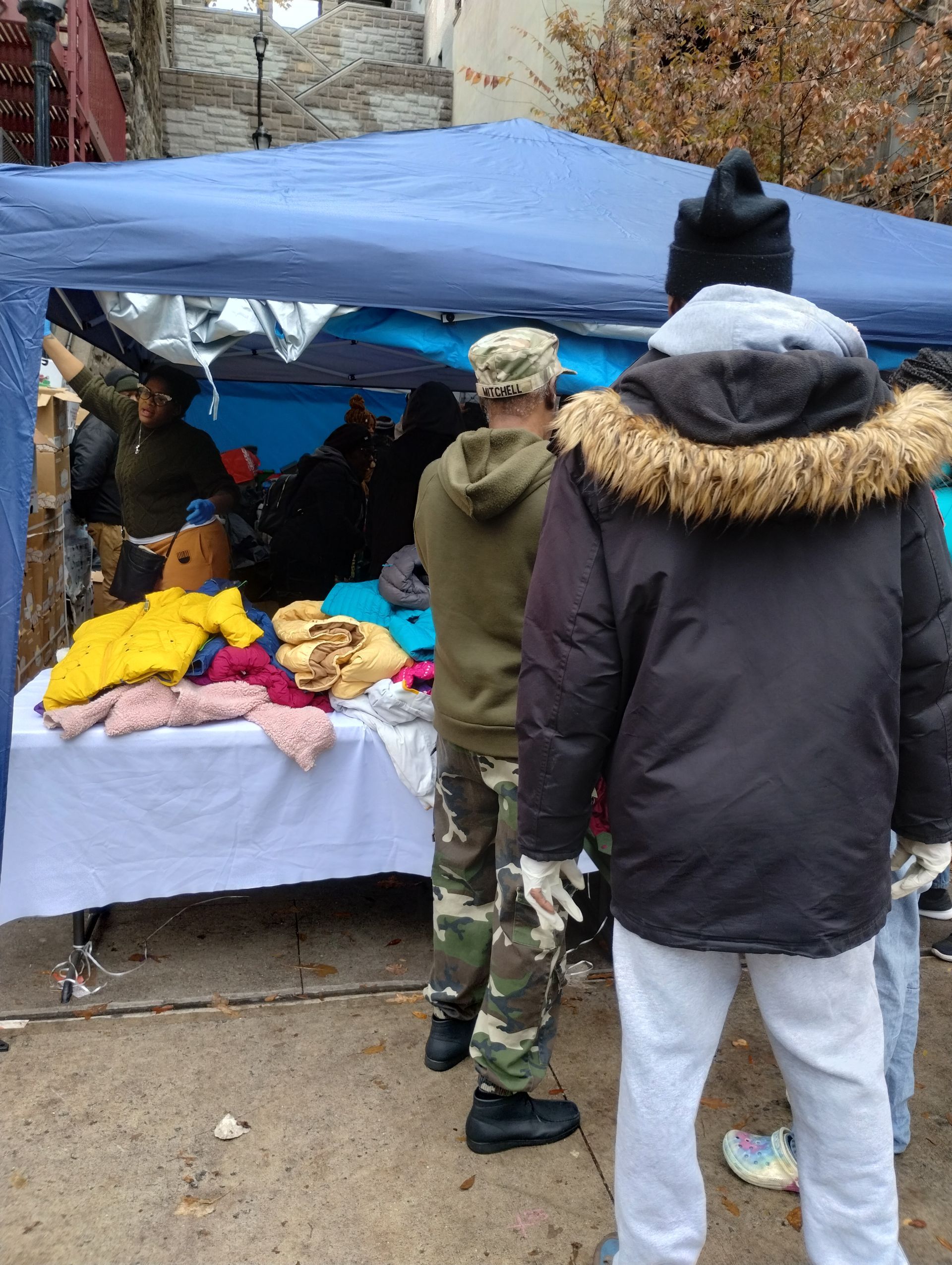 People browsing clothing displayed on a table under a blue tent outdoors.