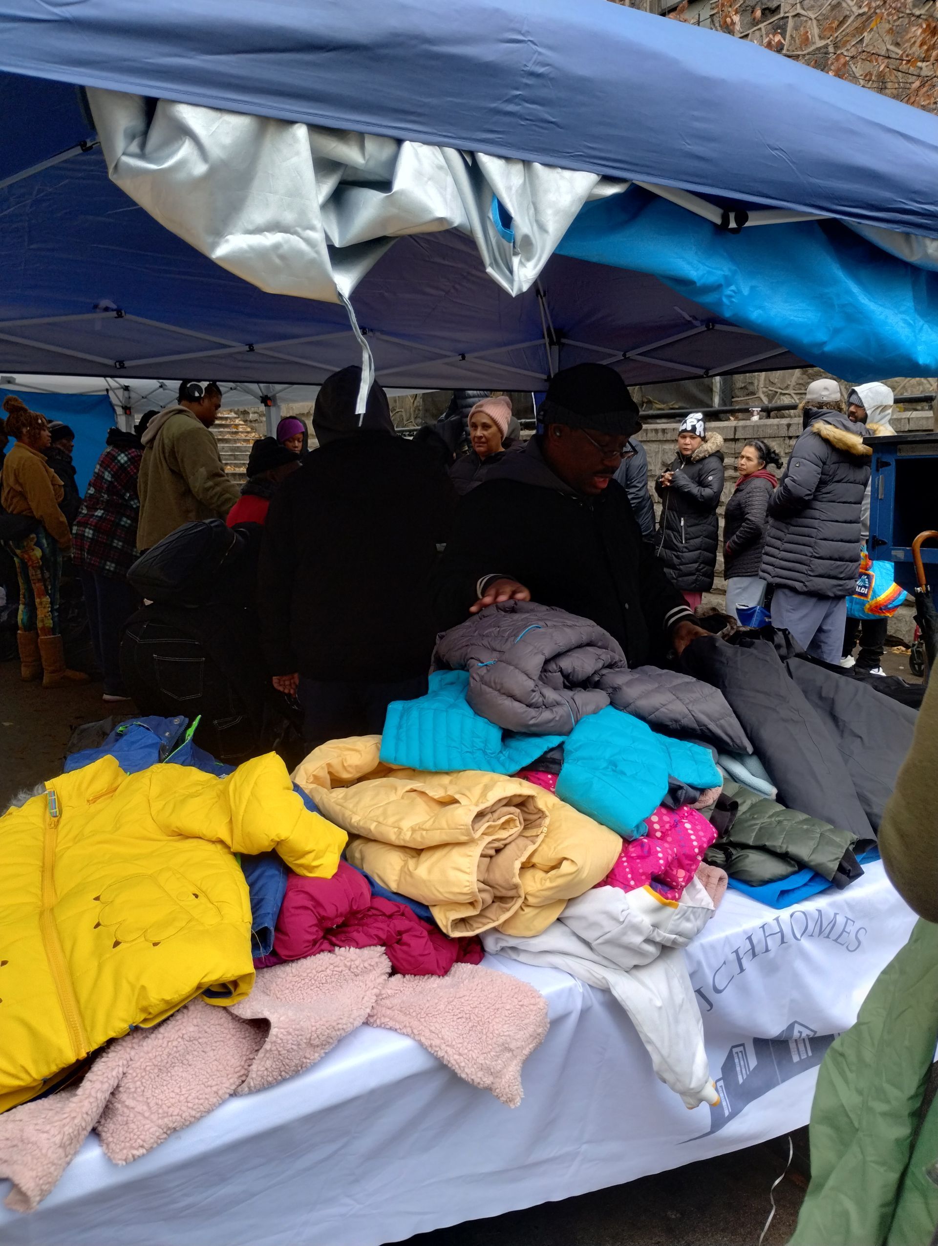 Outdoor market stall with jackets and clothing displayed. People browse under a blue tarp.