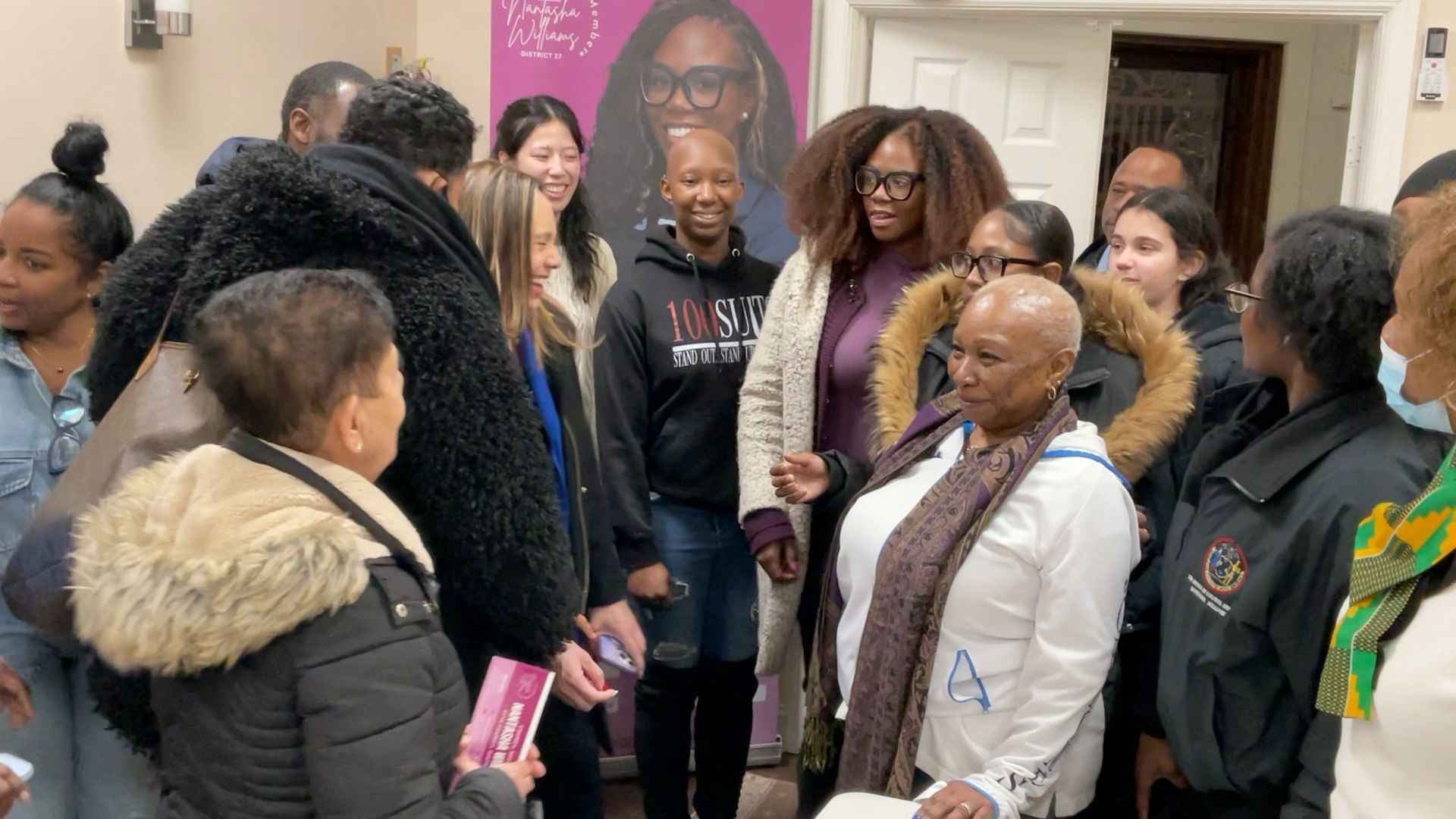 Group of people gathered indoors, smiling, possibly at an event. Purple banner in background.