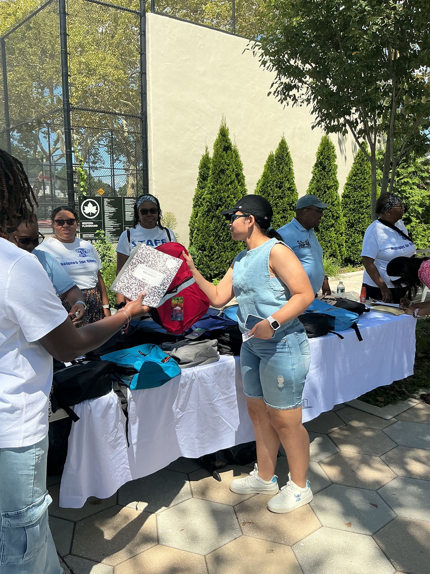 People browsing clothes and bags at an outdoor table under trees and a building wall