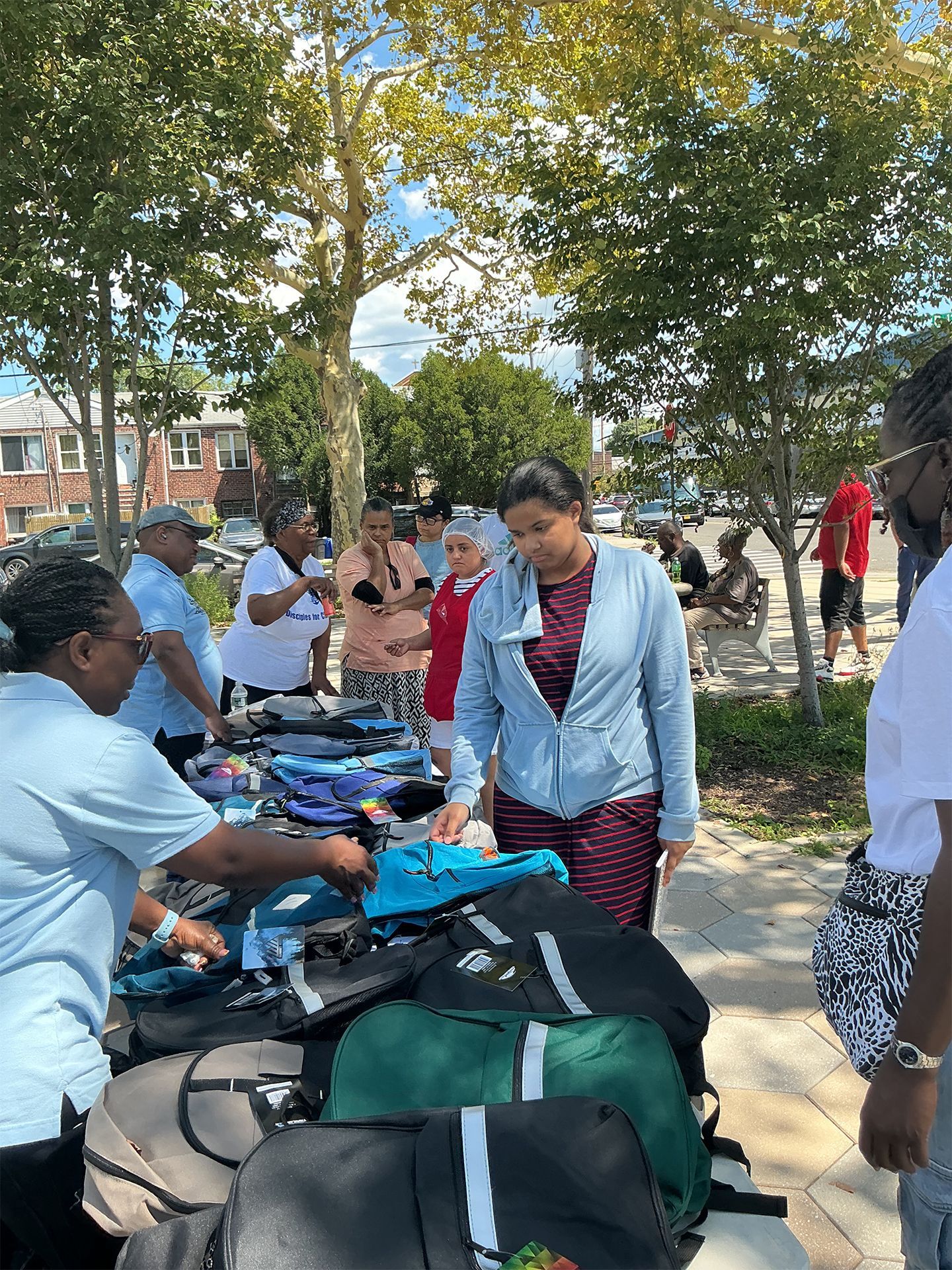 People gathered around a table outdoors under trees, chatting and handling bags and supplies.