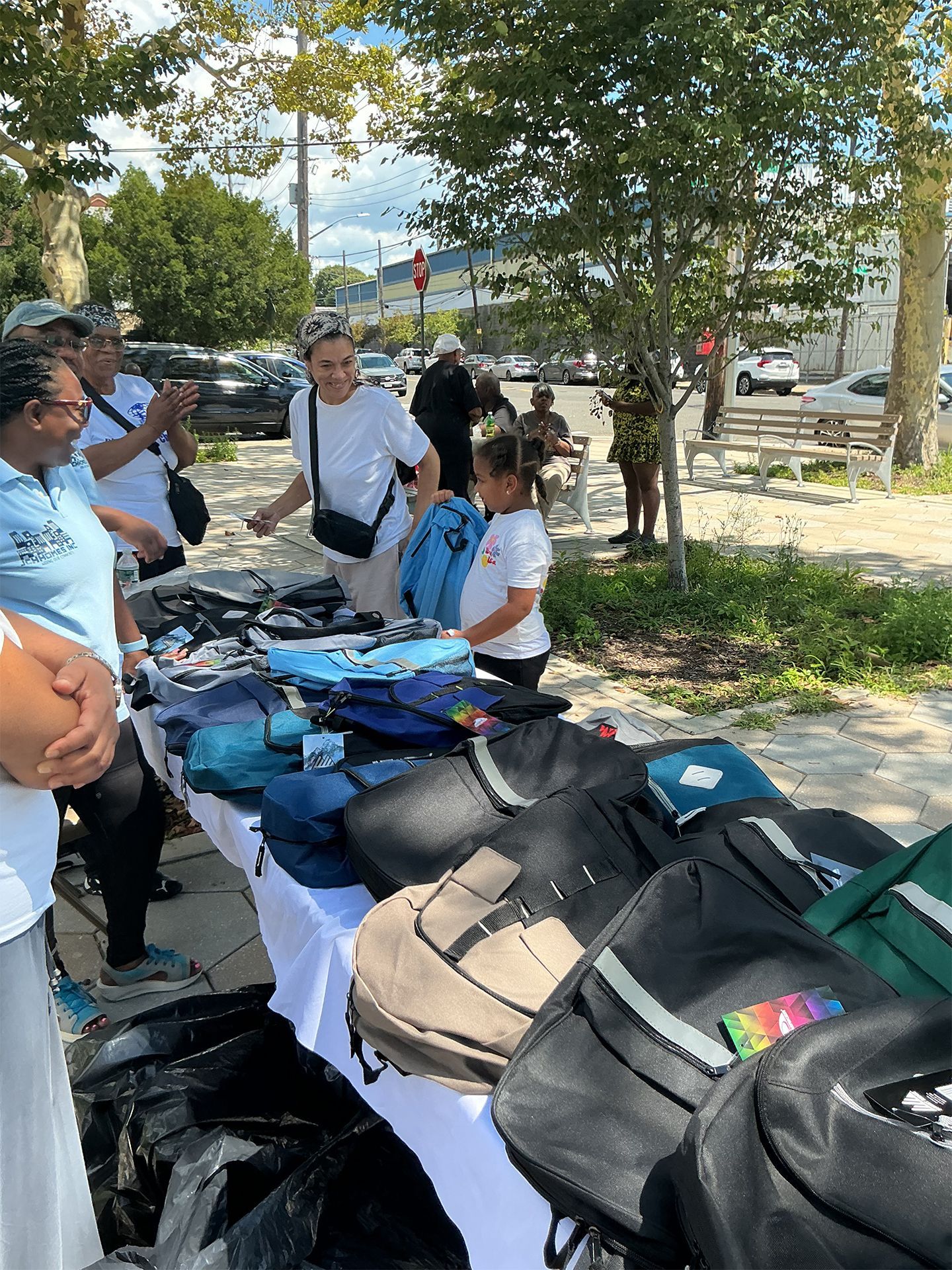 People browsing backpacks and bags laid out on a table at an outdoor market under trees.