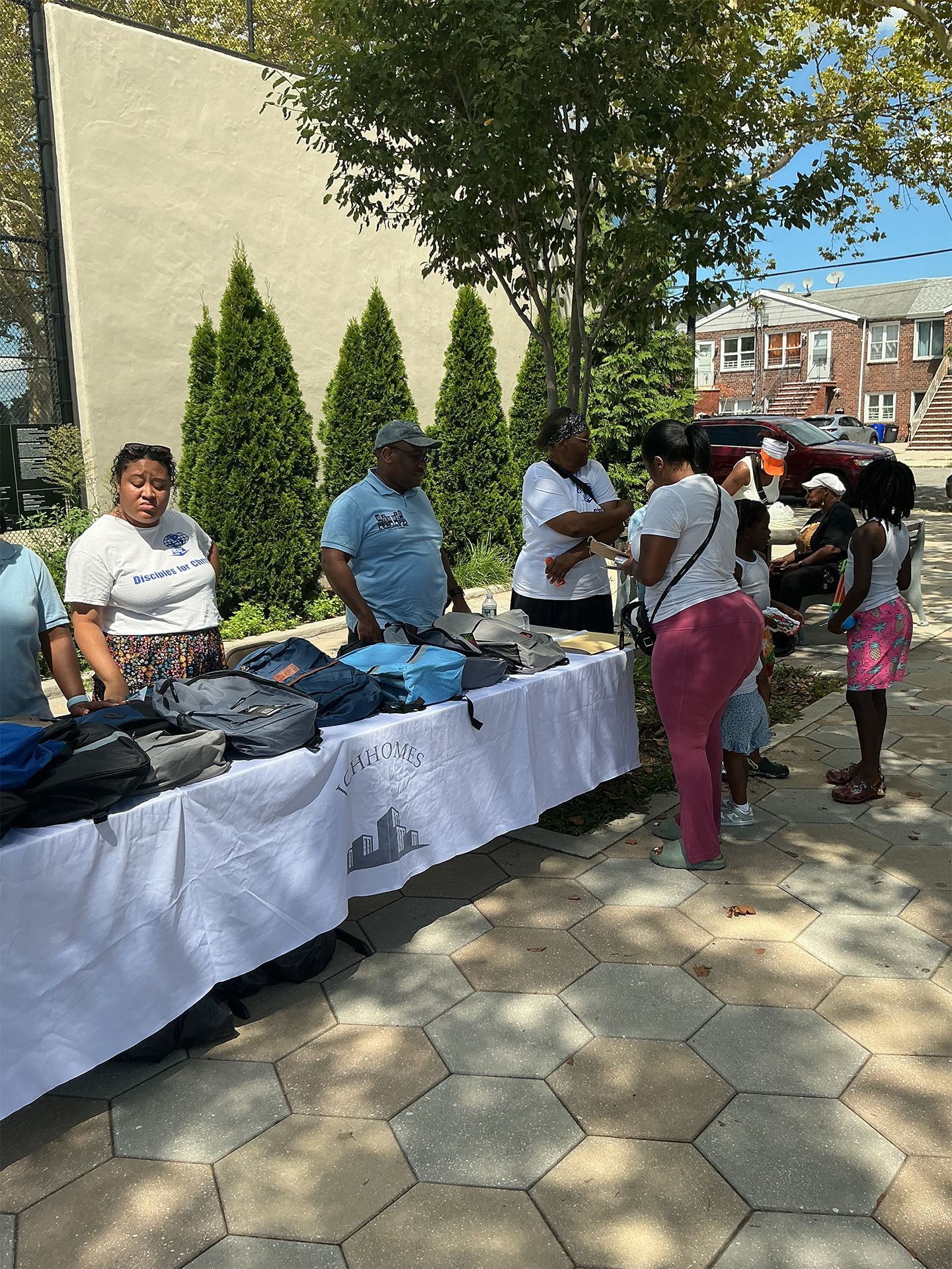 People handing out bags at a table outdoors on a sunny sidewalk by a building