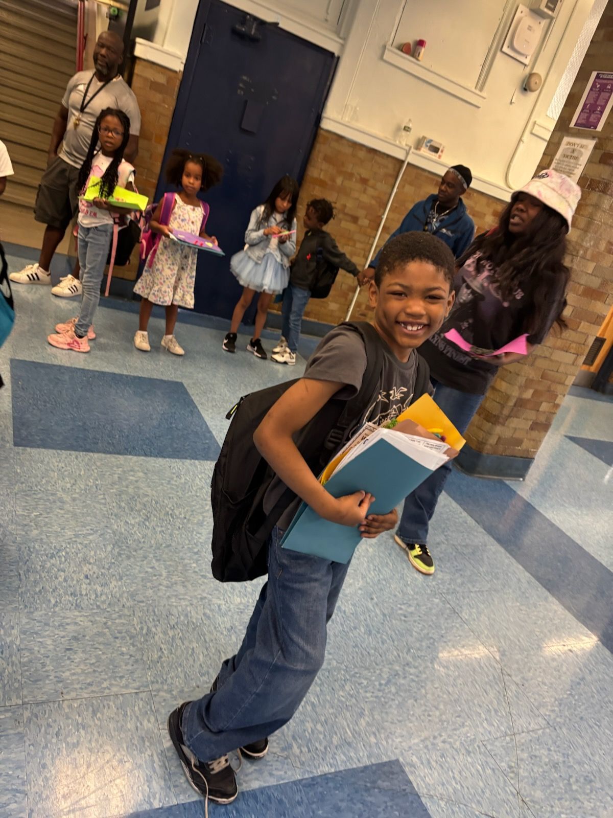 Boy in school hallway smiles, carrying books and backpack. Other students and adults are visible.