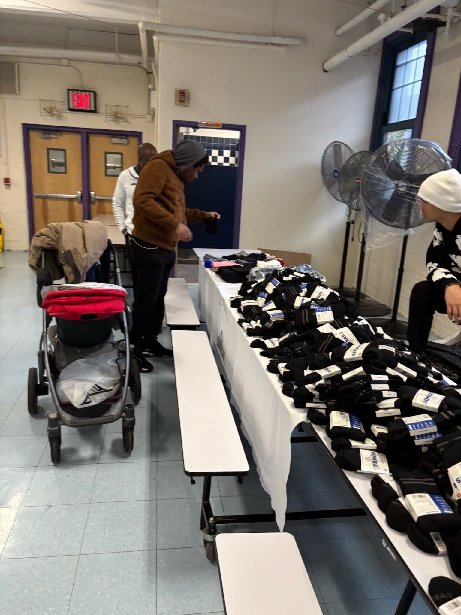 People at table with stacks of socks. A baby stroller is nearby. Indoors.