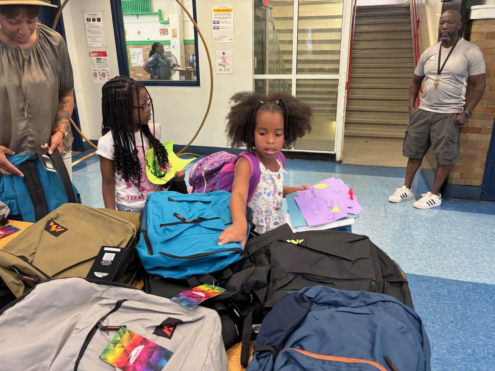 Children selecting backpacks from a table, likely at a school or community event. Adults nearby.