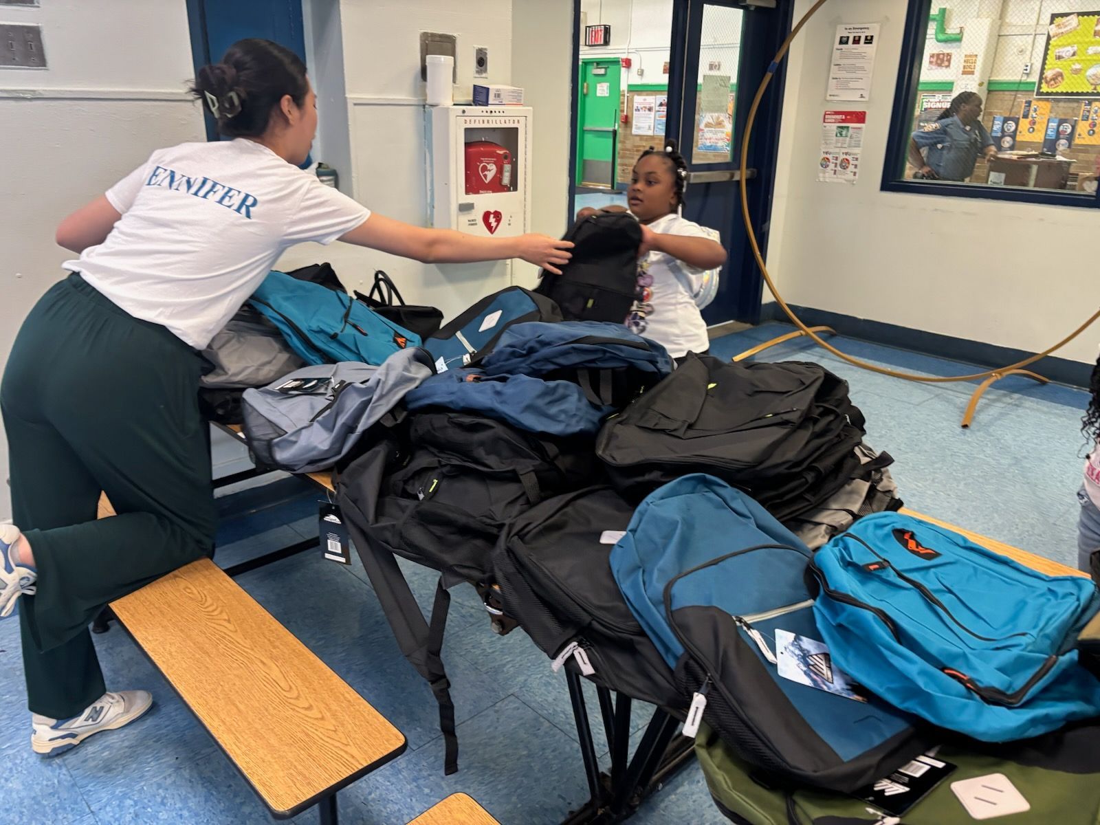 Two people organizing a table of backpacks indoors. One person hands another a black backpack.