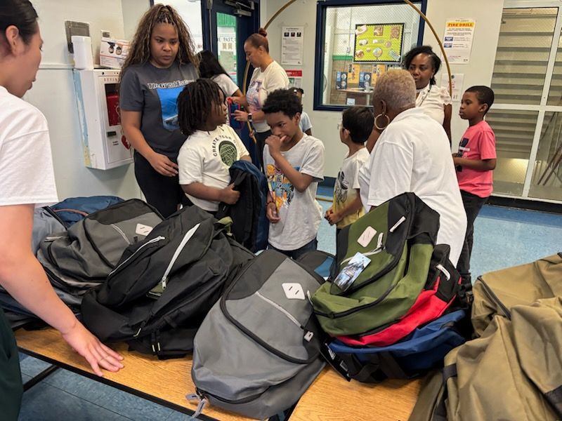 People and children choosing backpacks at a table in a brightly lit room.