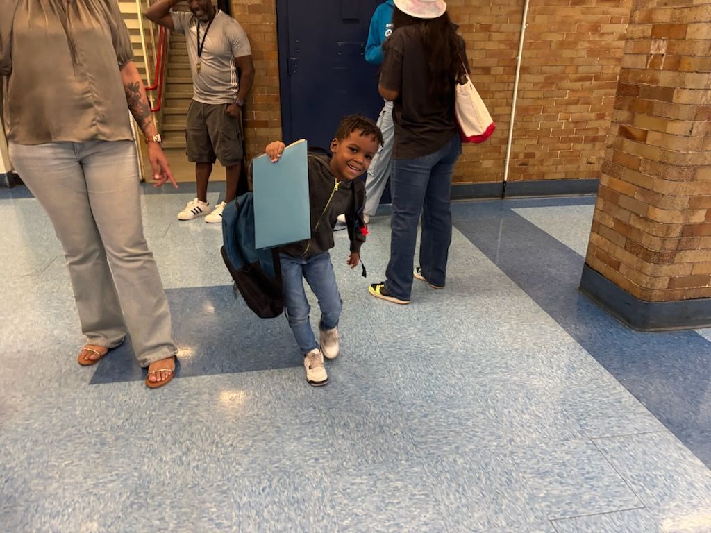 Young child in school hallway with backpack, holding paper, smiling.