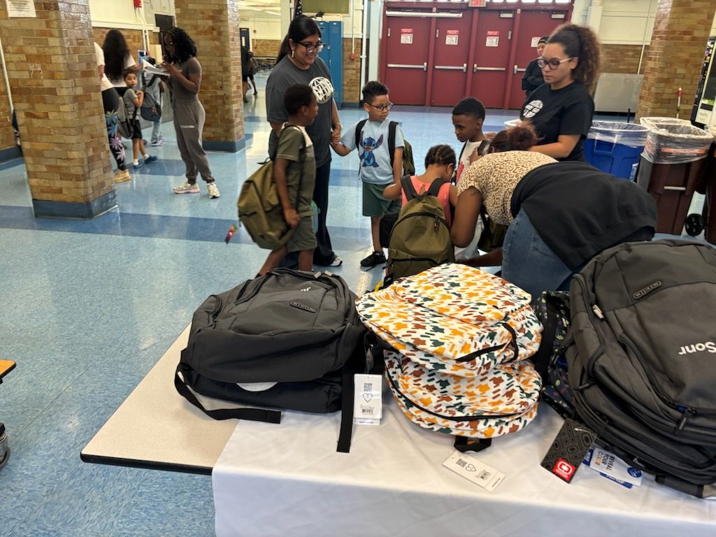 People selecting backpacks at a table, with children and adults in a school or community center.