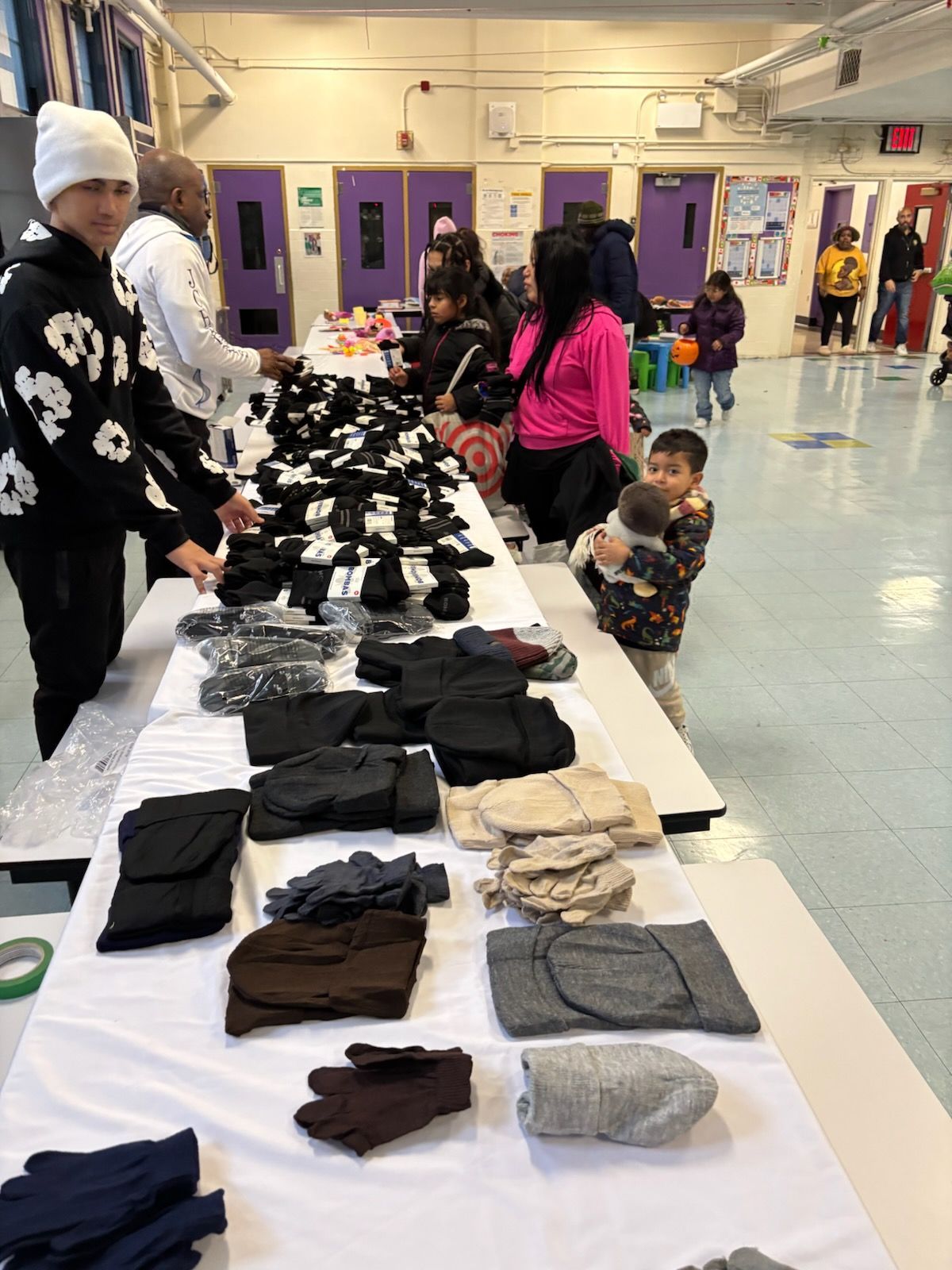 People at a table with stacks of black gloves and other winter items indoors; a child holds a baby.