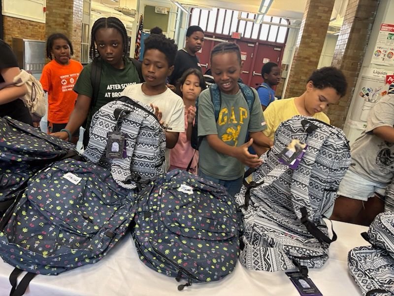 Children looking at backpacks on a table. Some are smiling, in a brightly lit room.