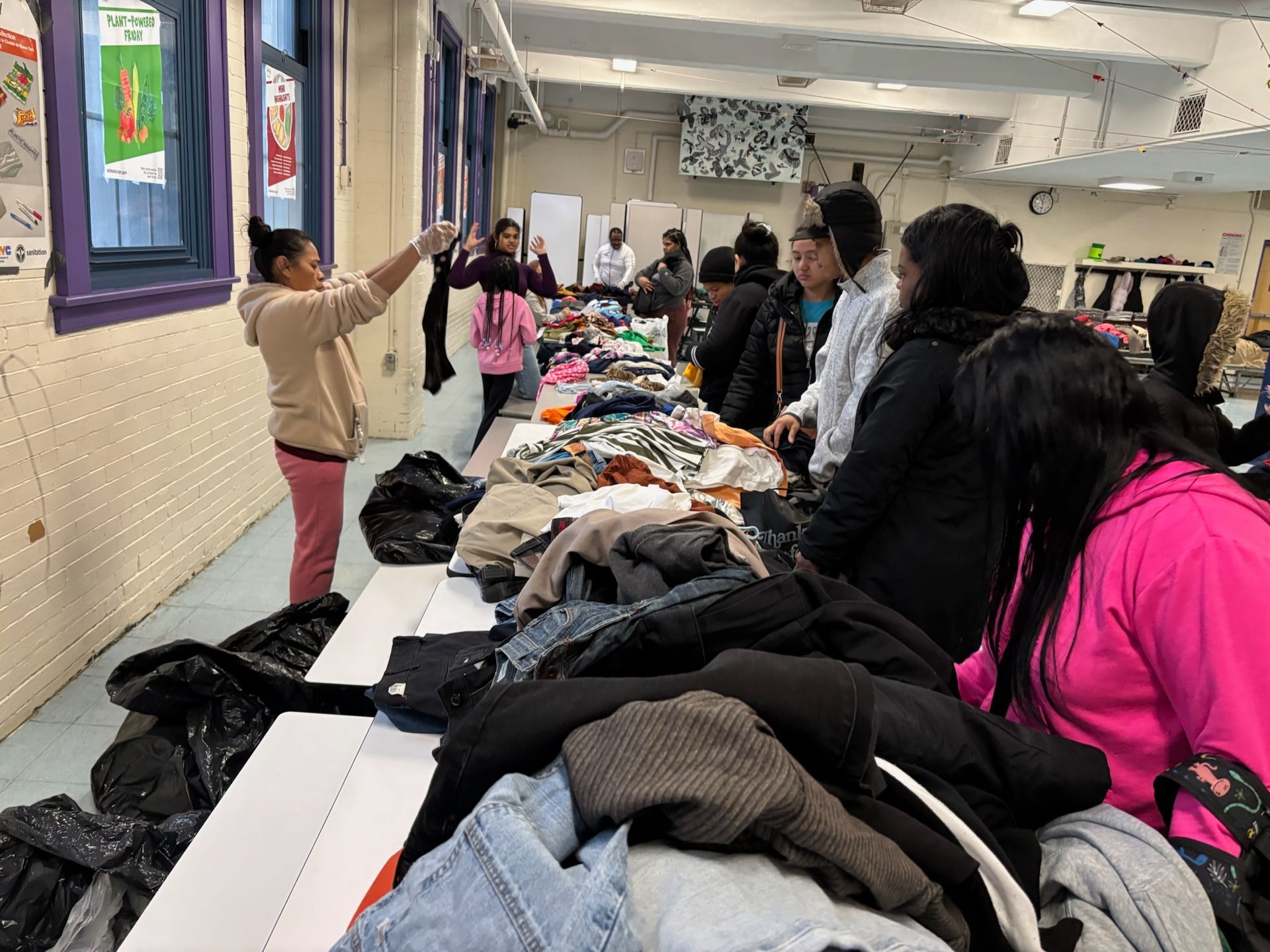 People browsing clothing laid out on tables in a room, one person holding up an item.