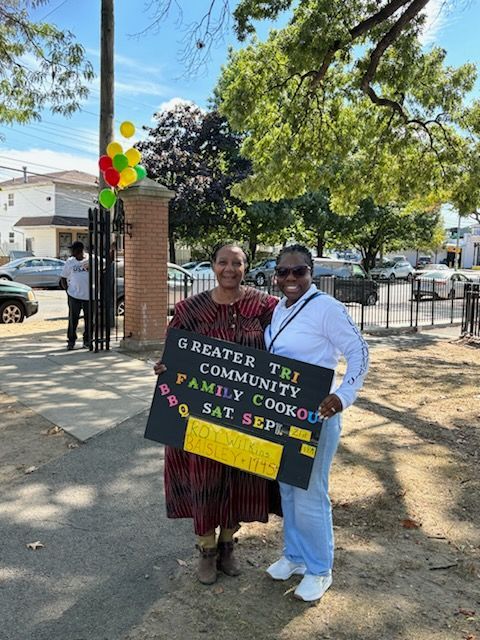 Two women holding a sign promoting a community cookout. Outdoor setting, colorful balloons in the background.