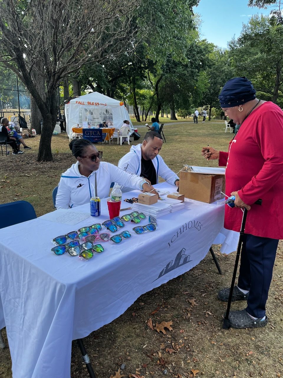Two people at a table selling sunglasses outdoors, one person buying. Green grass, trees, and tent visible.