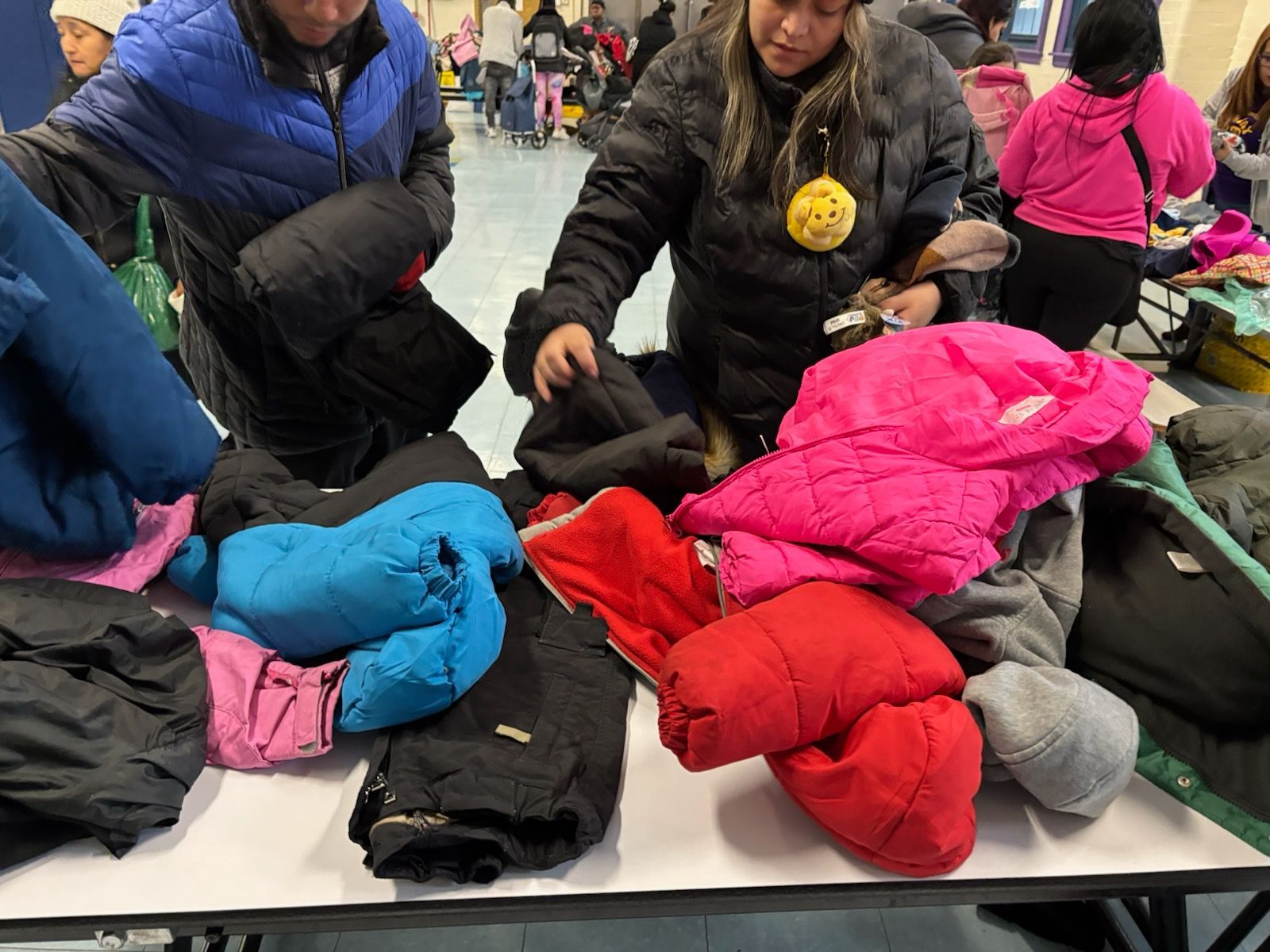 People sorting through jackets on a table indoors, likely for donation or distribution, with colorful coats visible.