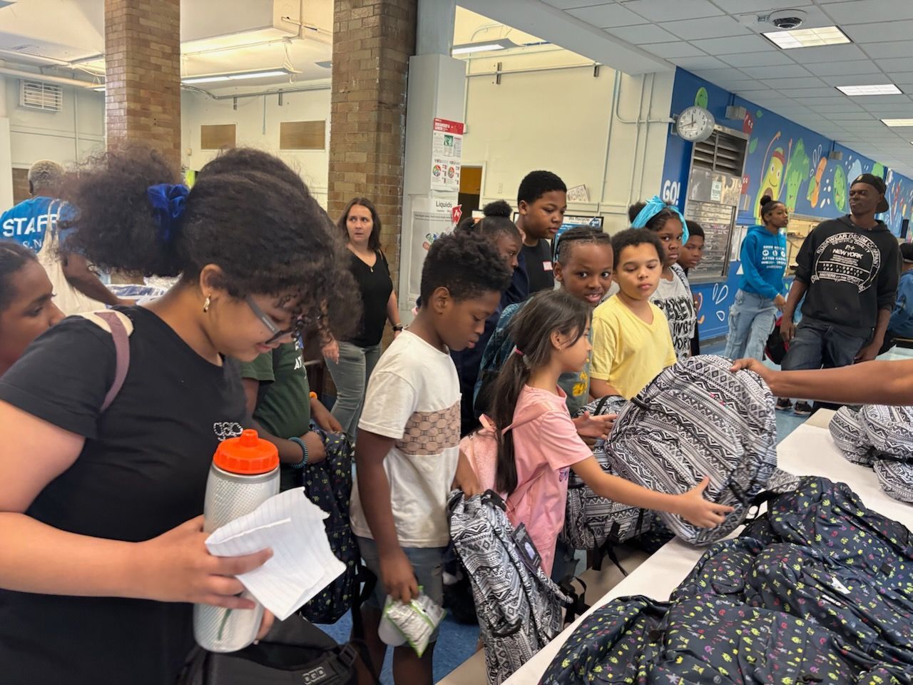 Children selecting backpacks from a table. People are in a room with a mural.