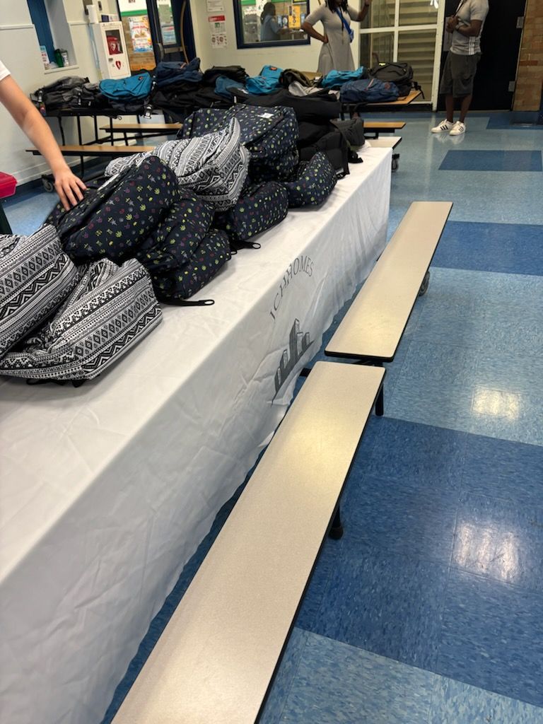 Backpacks piled on a covered table, in a school cafeteria with people in the background.