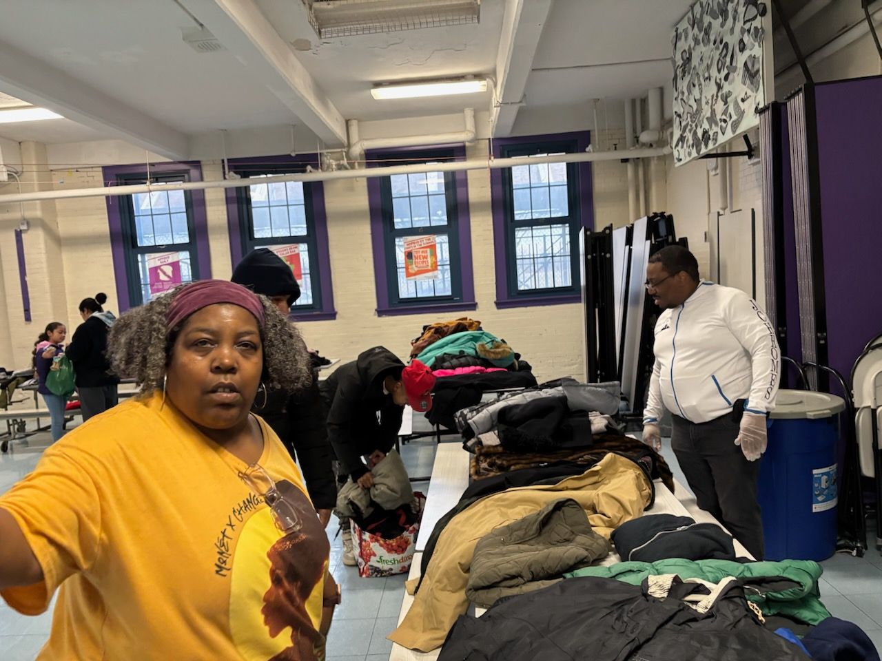 People sorting clothes in a room. Woman in yellow shirt looks at camera. Tables and windows are visible.
