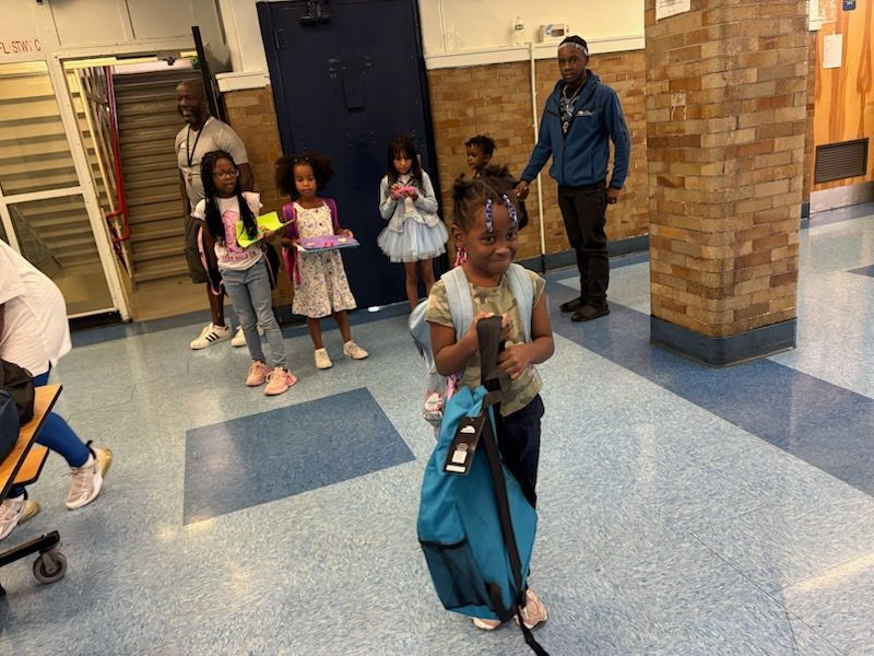 Children with backpacks in a school hallway, accompanied by adults.