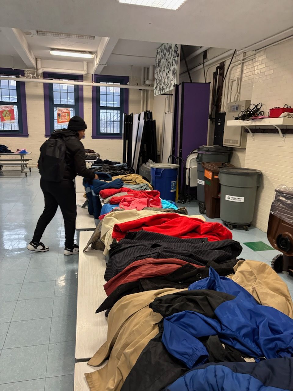 Person selecting clothing laid out on tables in a room with a window, trash cans, and other items.