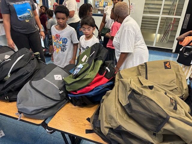 Backpacks on a table with children and adults gathered nearby, possibly at a school or community event.