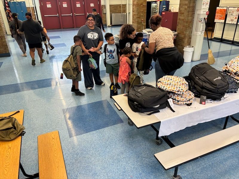 People selecting backpacks from a table in a school hallway. Several children and adults are present.