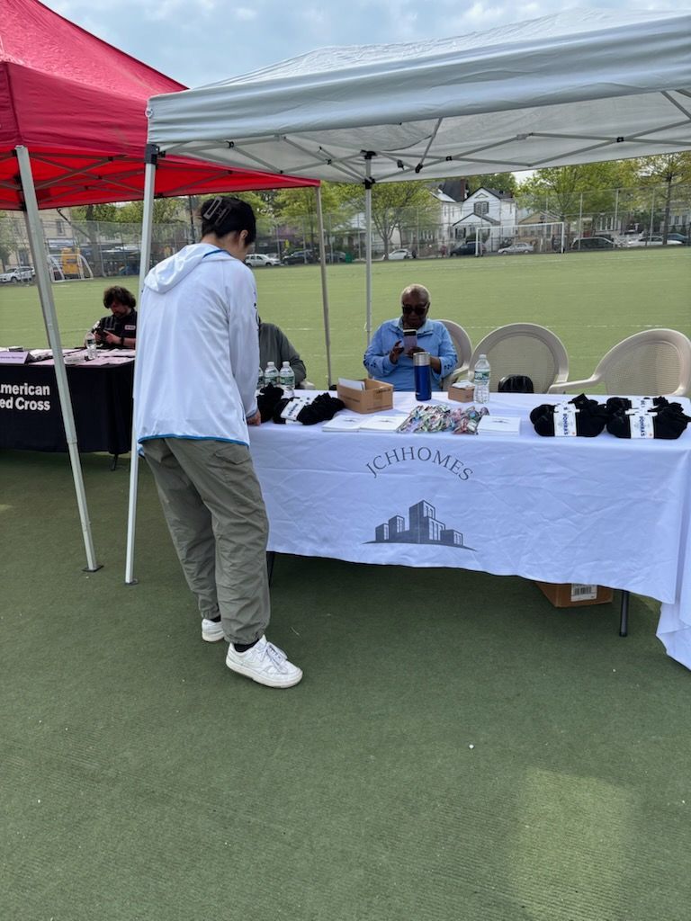 Person looking at items on a table under a canopy. Other people are behind the table. Outdoor setting.