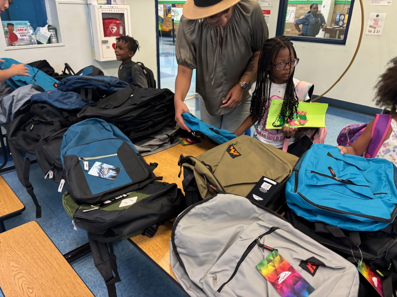 People selecting backpacks from a table filled with various colors in a school setting.