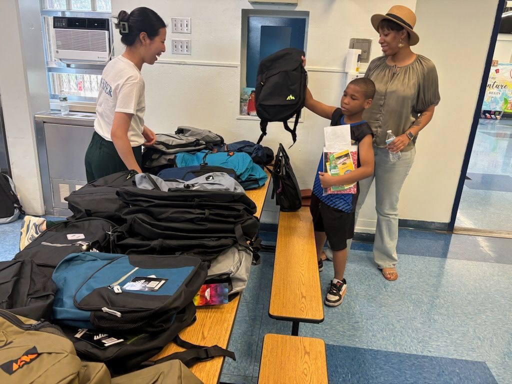 Person giving a backpack to a child; table with more backpacks, another person watching. Indoors.
