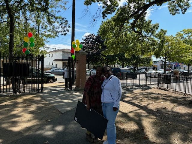 Two people stand at park entrance, holding a dark object. Balloons in yellow, green, red are tied to the gate.