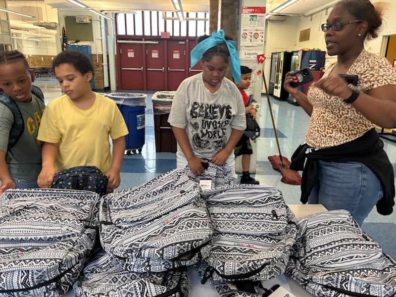 Children and adult selecting backpacks. A table displays many backpacks with black and white designs inside a building.