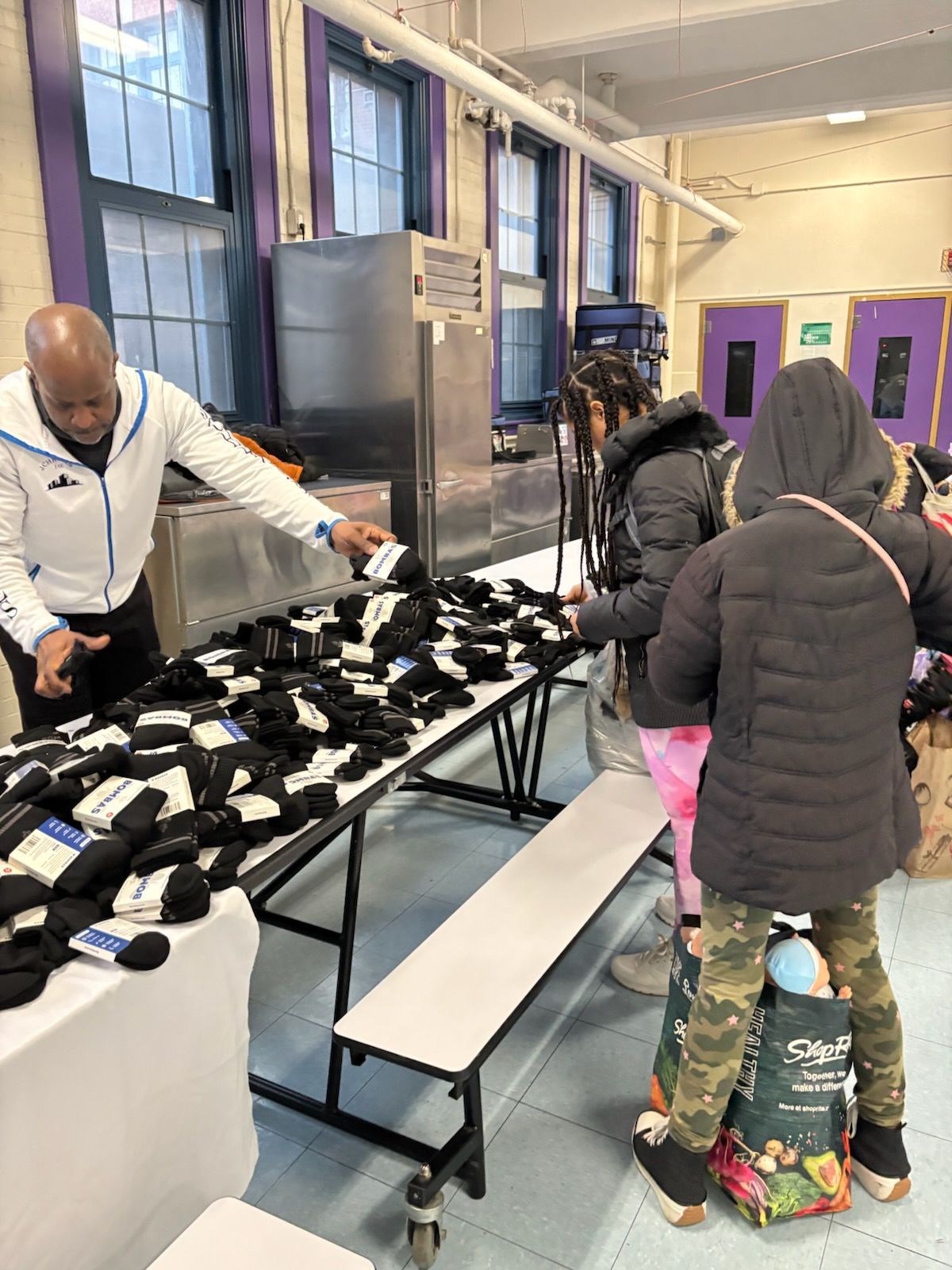 Person distributing black socks at a table to two children. A stainless steel appliance and windows are visible.