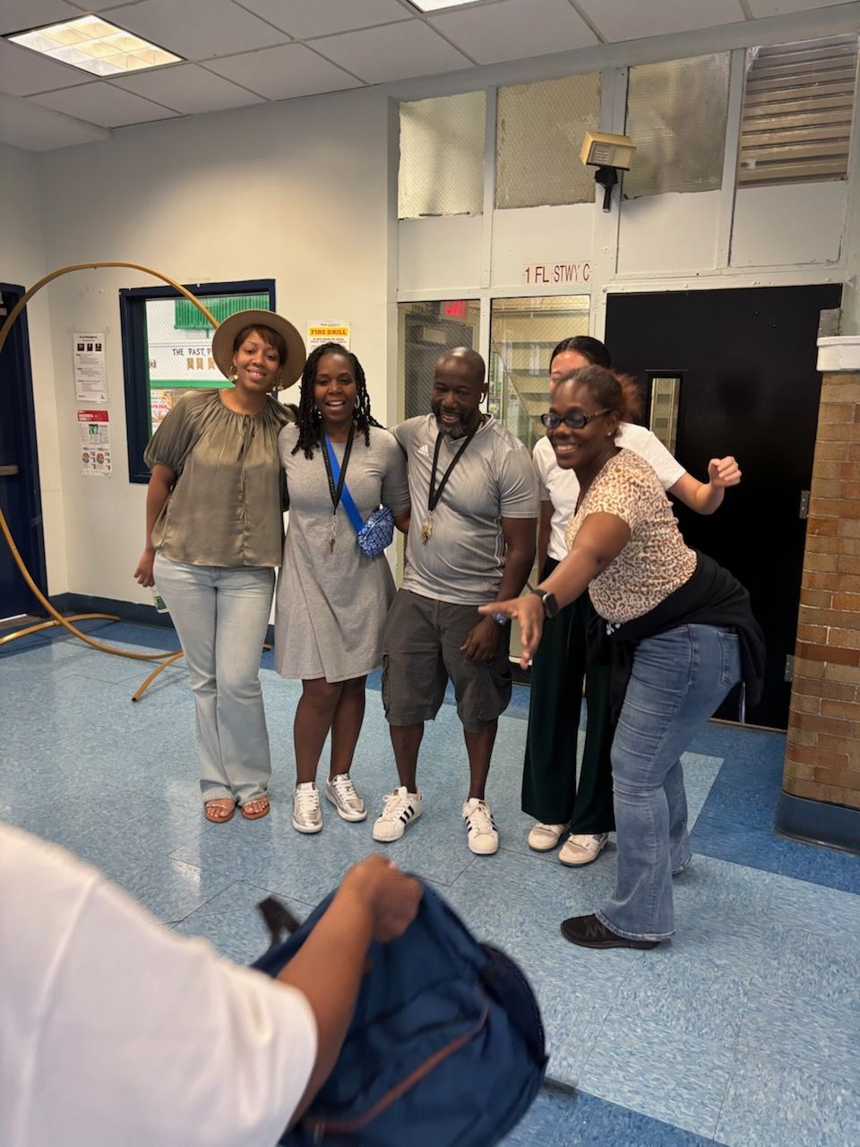 Group of people posing for a photo indoors. Two women and a man smile, while another woman pretends to take photo, and one person holds a backpack.
