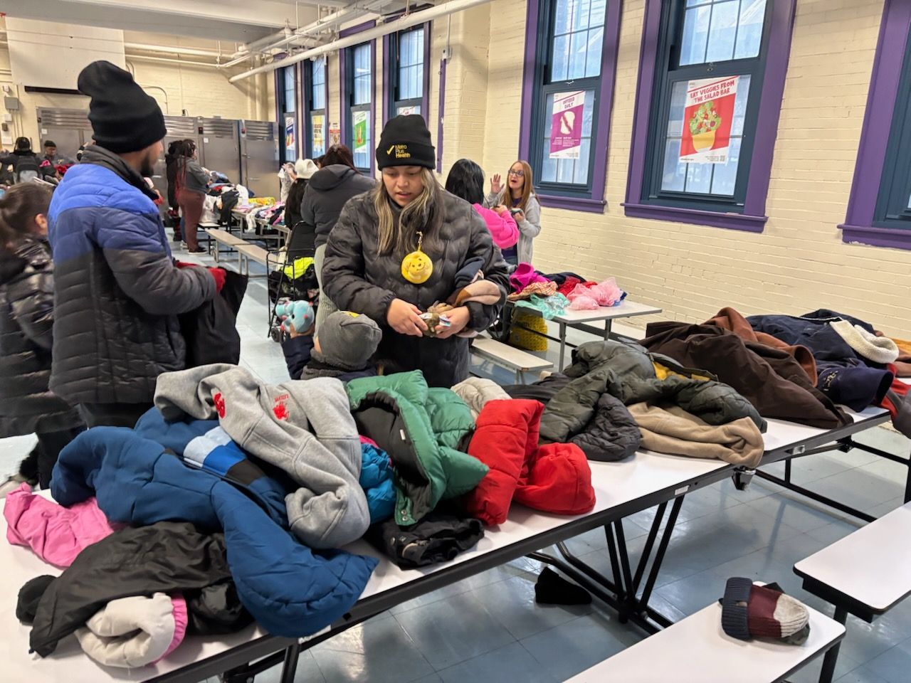 People selecting coats from a table in a school cafeteria. Several coats of various colors are spread out on the table.