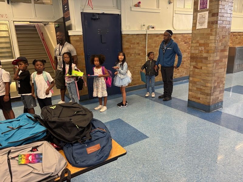 Children with adults in a school hallway with backpacks on the floor.
