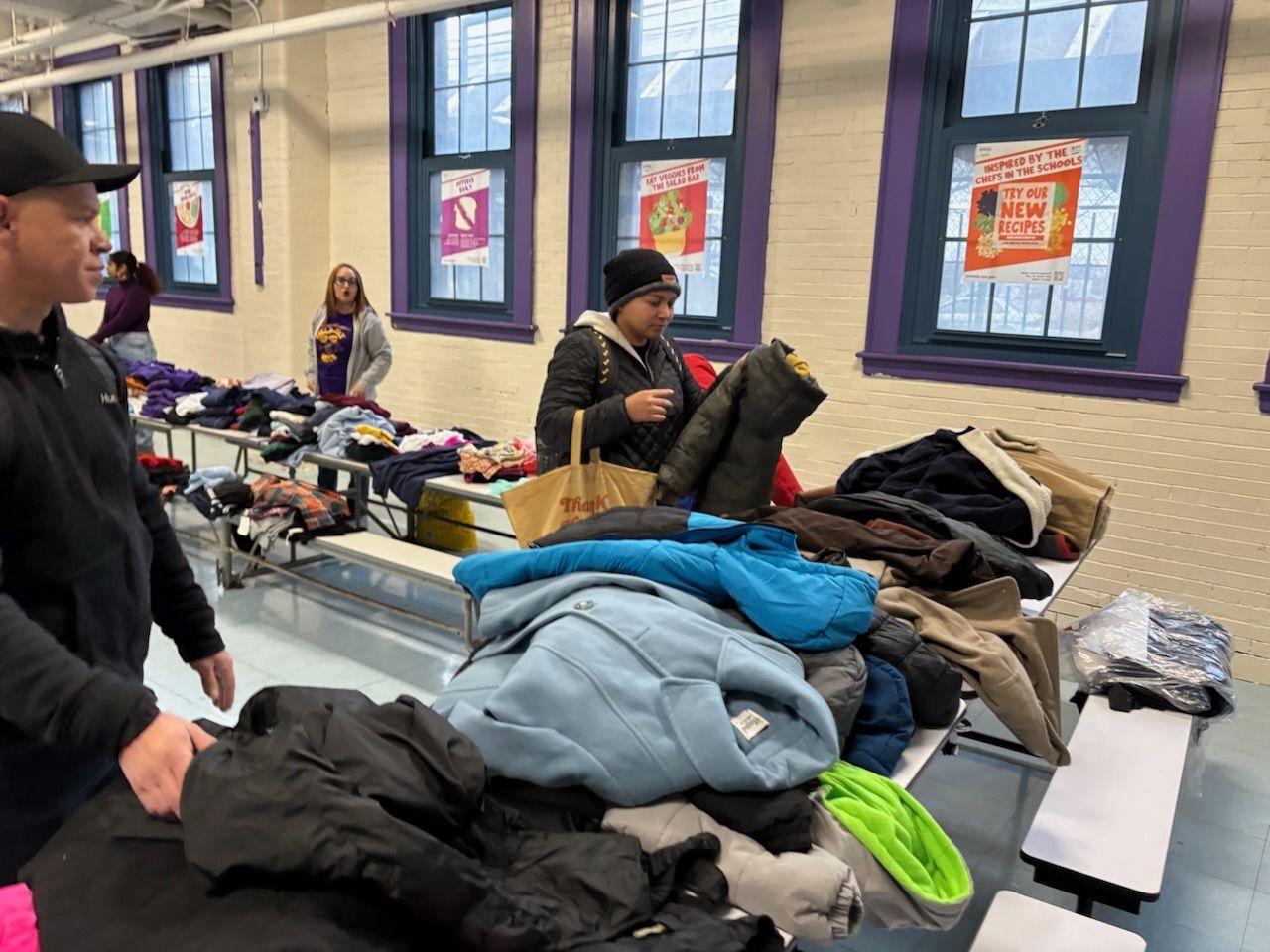 People selecting clothes from tables in a brightly lit room with windows.