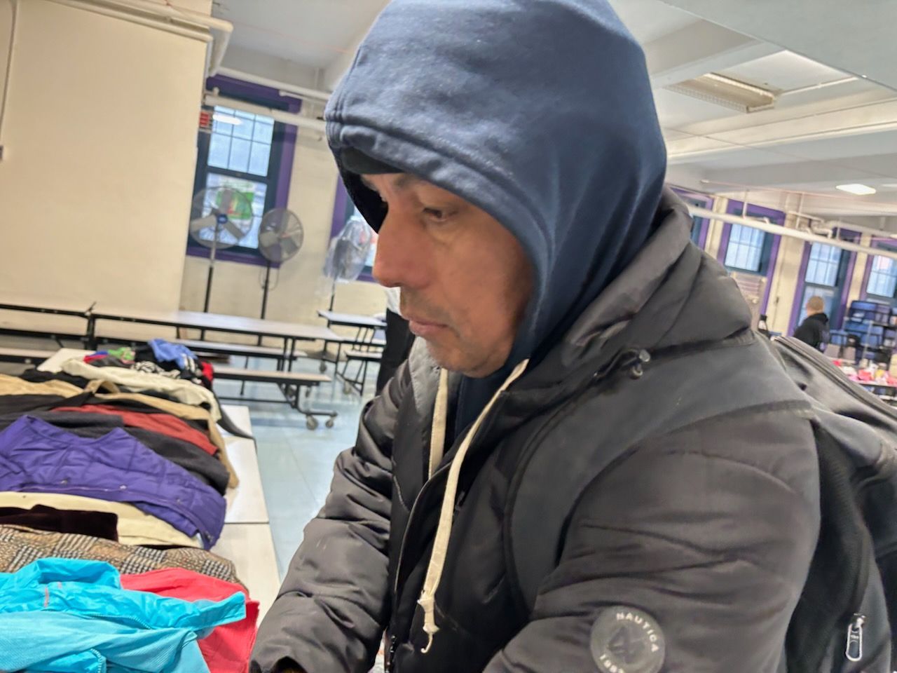 Man in a dark hooded jacket looking at clothes on a table inside a building with a beige and purple interior.