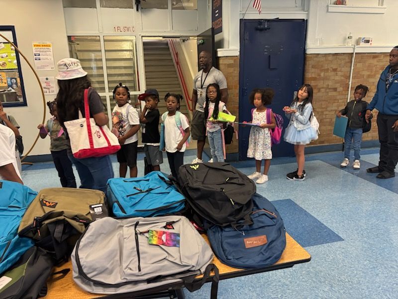 Children receiving backpacks in a school hallway. Several adults are present.