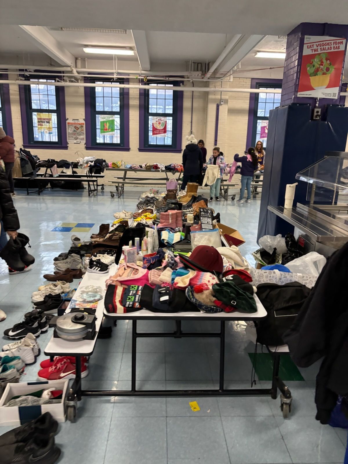 A table with shoes and clothes set up in a school cafeteria, people browsing items.